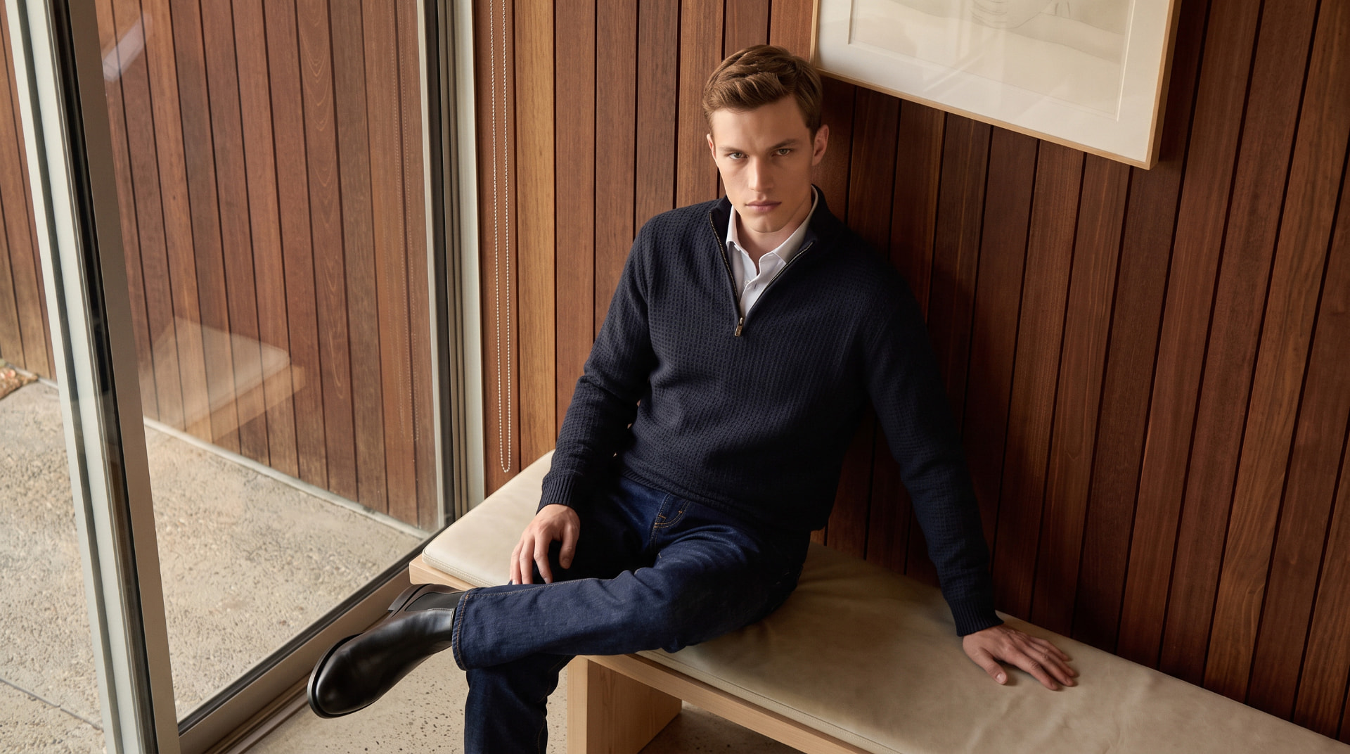 Man in a navy quarter-zip sweater sitting on a bench beside a wood paneled wall