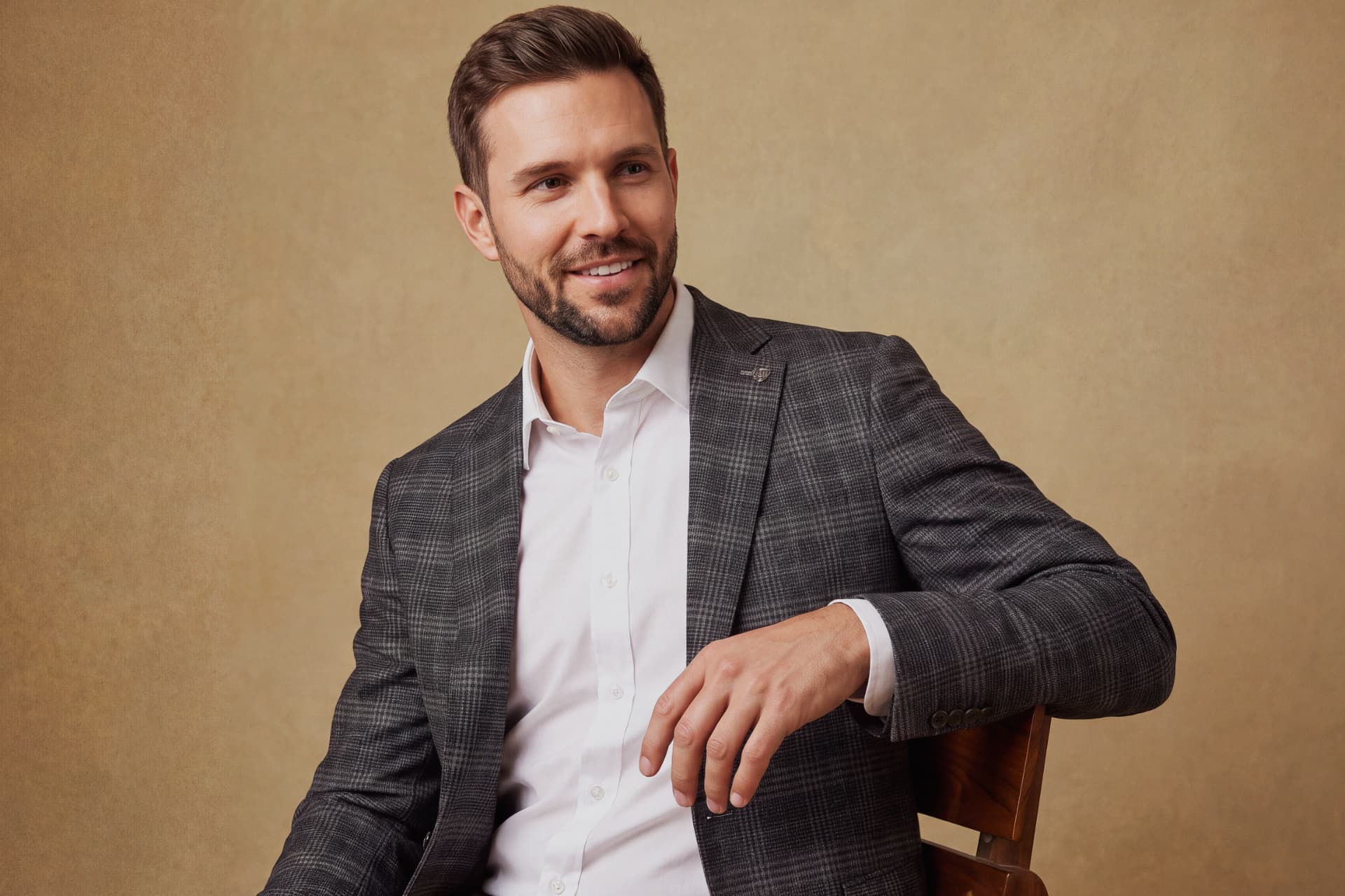 Man in a grey plaid Tom Baine blazer over a white dress shirt, seated against a warm tan background